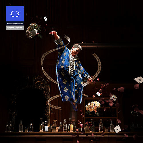 A man in a blue suit hovering over a bar with empty bottles on it. He is surrounded by cards and flowers floating around him.